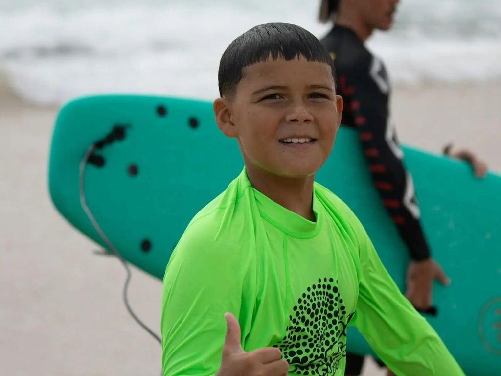 Boy gives thumbs up after surf lesson, green surfboard in background