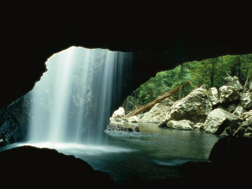 A sheet of white water, backlit by sunlight, falls into a dark rockpool,  underneath a rock arch.