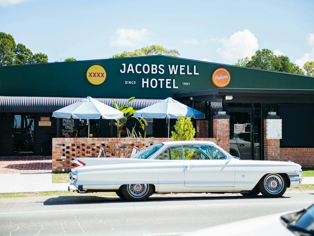 A vintage white Cadillac parked in front of the dark green Jacobs Well Hotel building on a sunny day
