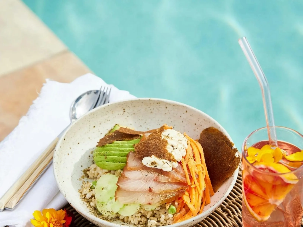 Bowl of colourful vegetables and fish with an iced tea overlooking pool