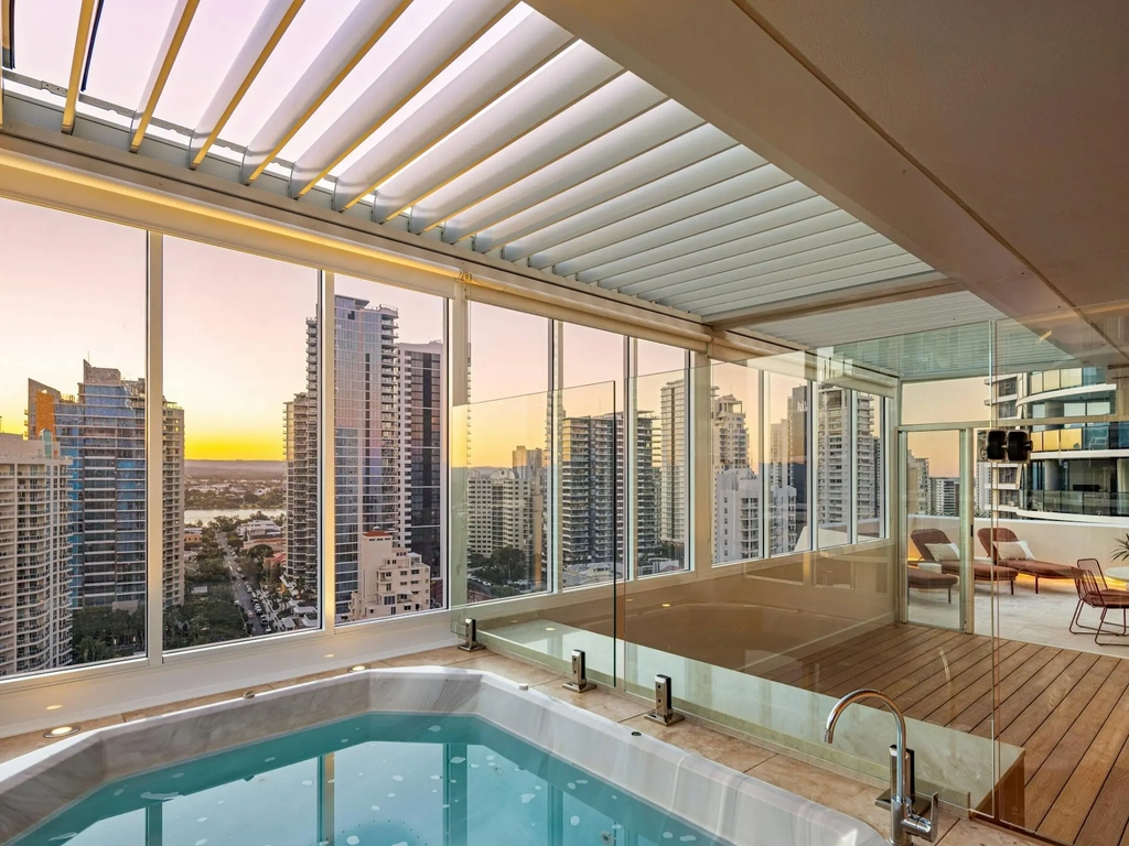 Private rooftop spa enclosed in glass under a louvred roof, city towers at sunset