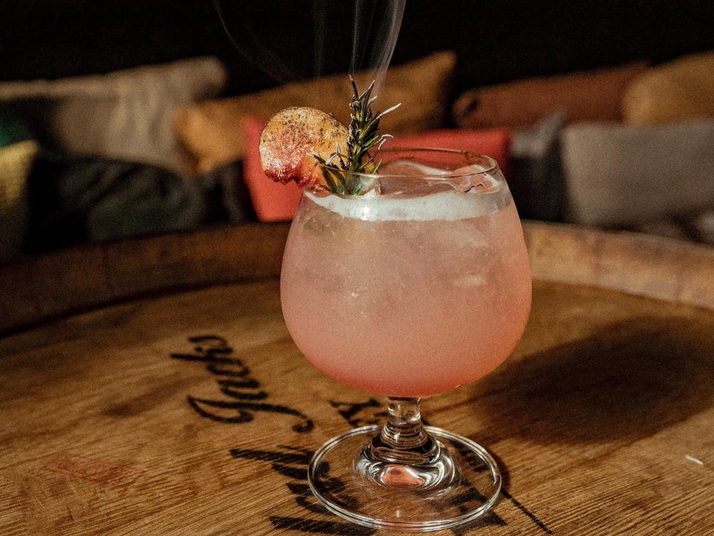 A photo of a pink cocktail in a balloon glass with a burnt rosemary leaf and dehydrated fruit.