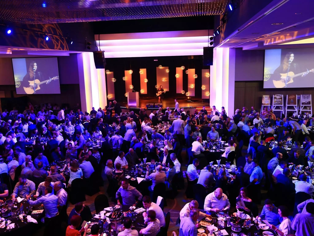 A crowd of people attending an event, seated at tables with a musician on stage