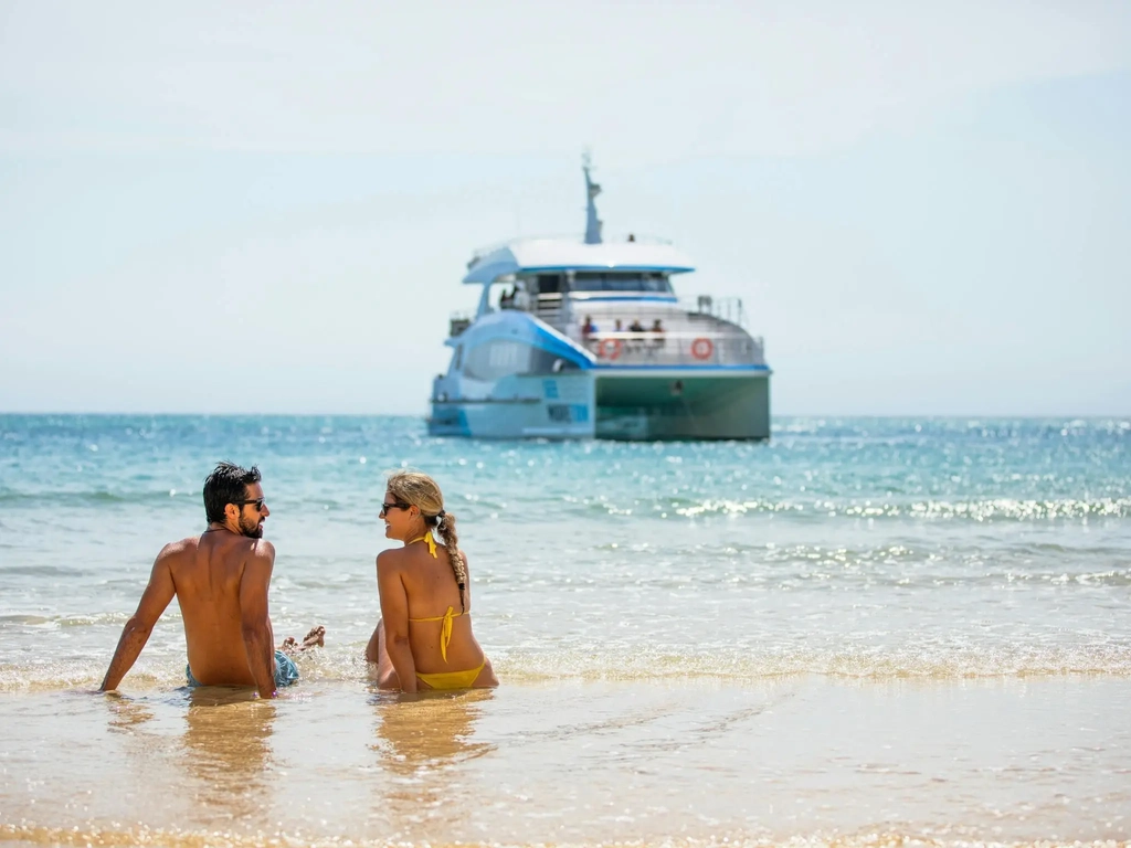 Man and woman in swim wear sit on beach with boat in distance.