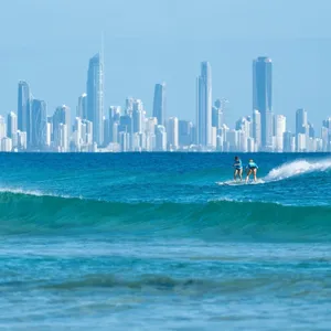 Two women surfing in blue ocean with city skyline in background.