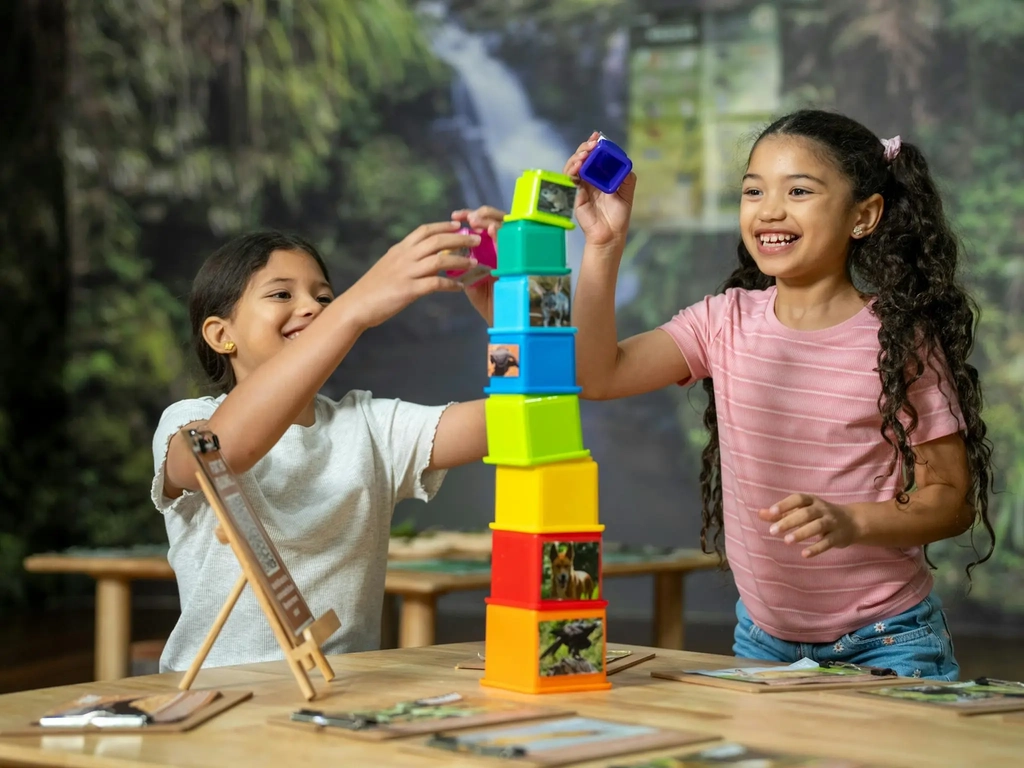 Two smiling girls stack colourful plastic bocks to form a tapered tower in a rainforest themed room.