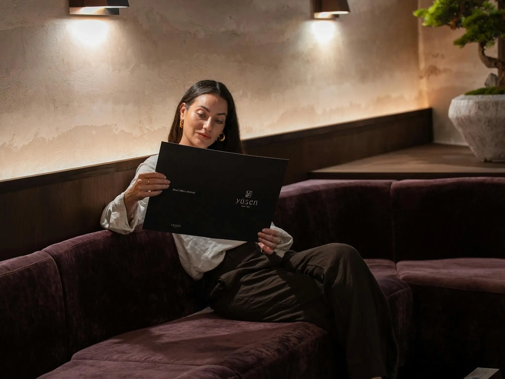 Relaxed guest reviewing the treatment menu booklet beside a bonsai tree and warm wall lighting.