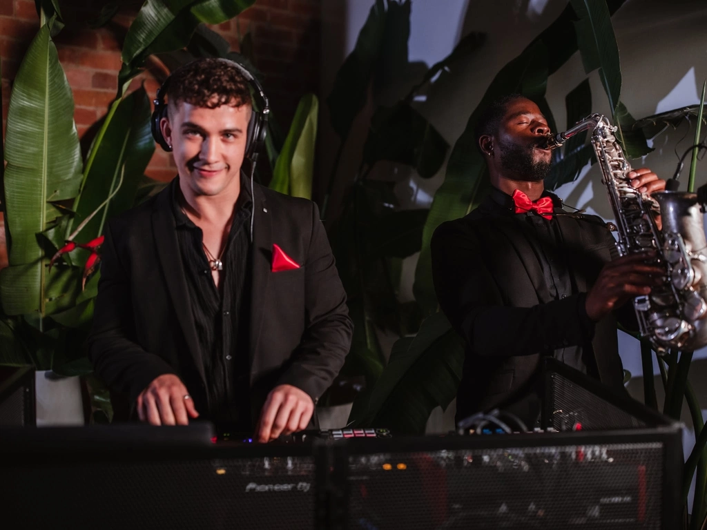 A DJ has his hands on the DJ decks while a saxophonist plays next to him,