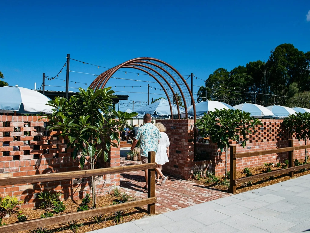 A couple walks through a brick archway entrance into a sunlit outdoor patio area with umbrellas