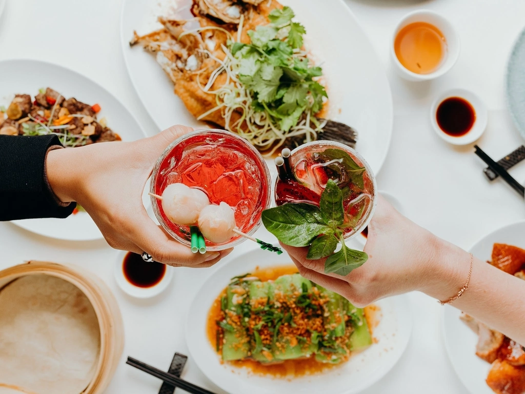 Two people hold two drinks above a spread of food.