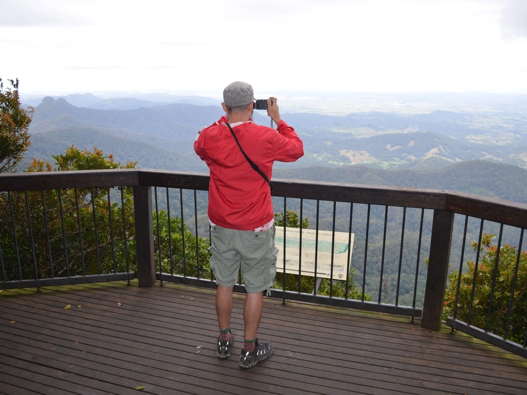 Man in red jacket looks thorugh binoculars at a lookout with views of mountains and valley.