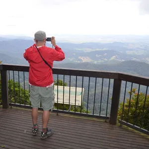 Man in red jacket looks thorugh binoculars at a lookout with views of mountains and valley.