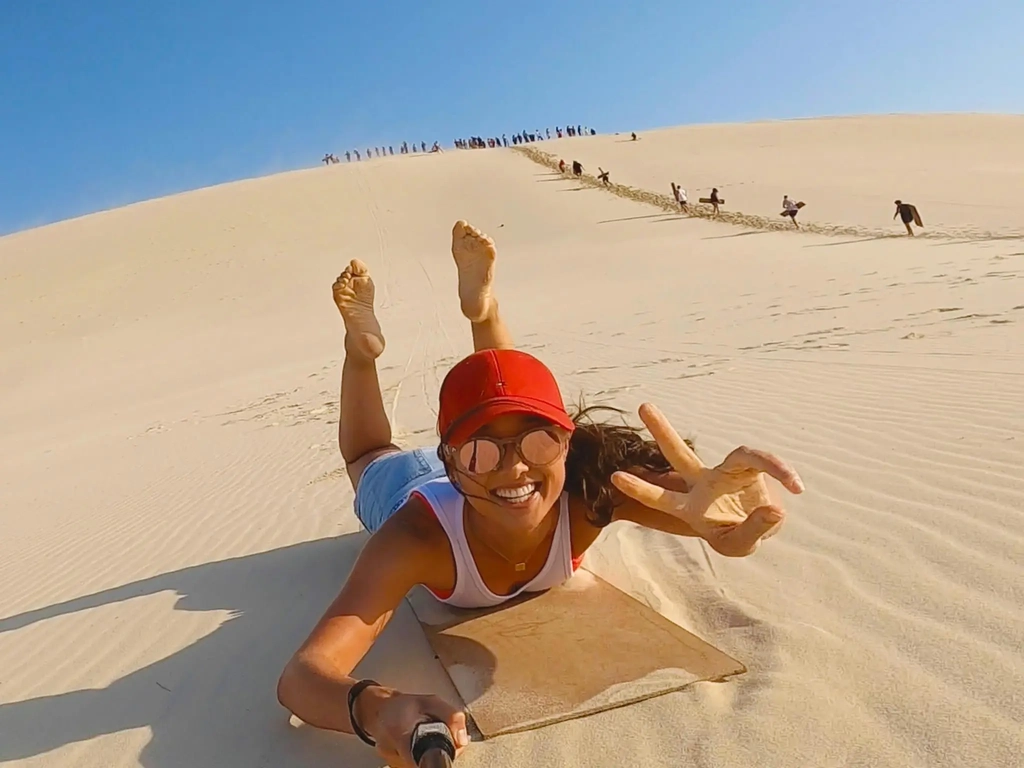 Women on a sandboard riding down a sand due, holding a gopro in one hand