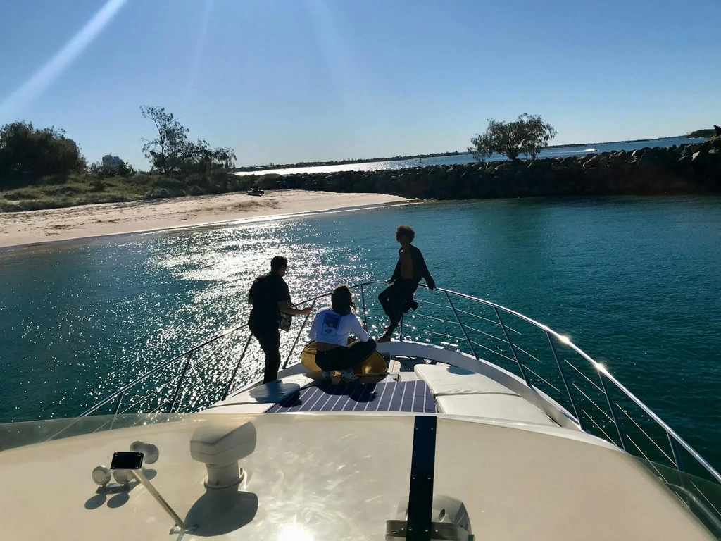 Three people enjoying the bow of a private yacht anchored near a sandy beach on the Gold Coast.