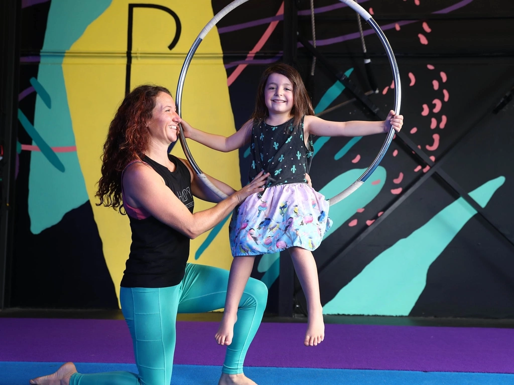 Kids Aerial Classes - Teacher is supporting a  young girl who is sitting on an Aerial hoop.