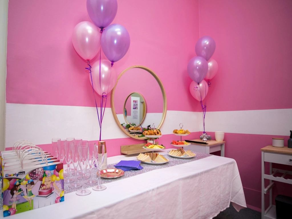 A long table with a white table cloth with pink and purple balloons and party bags
