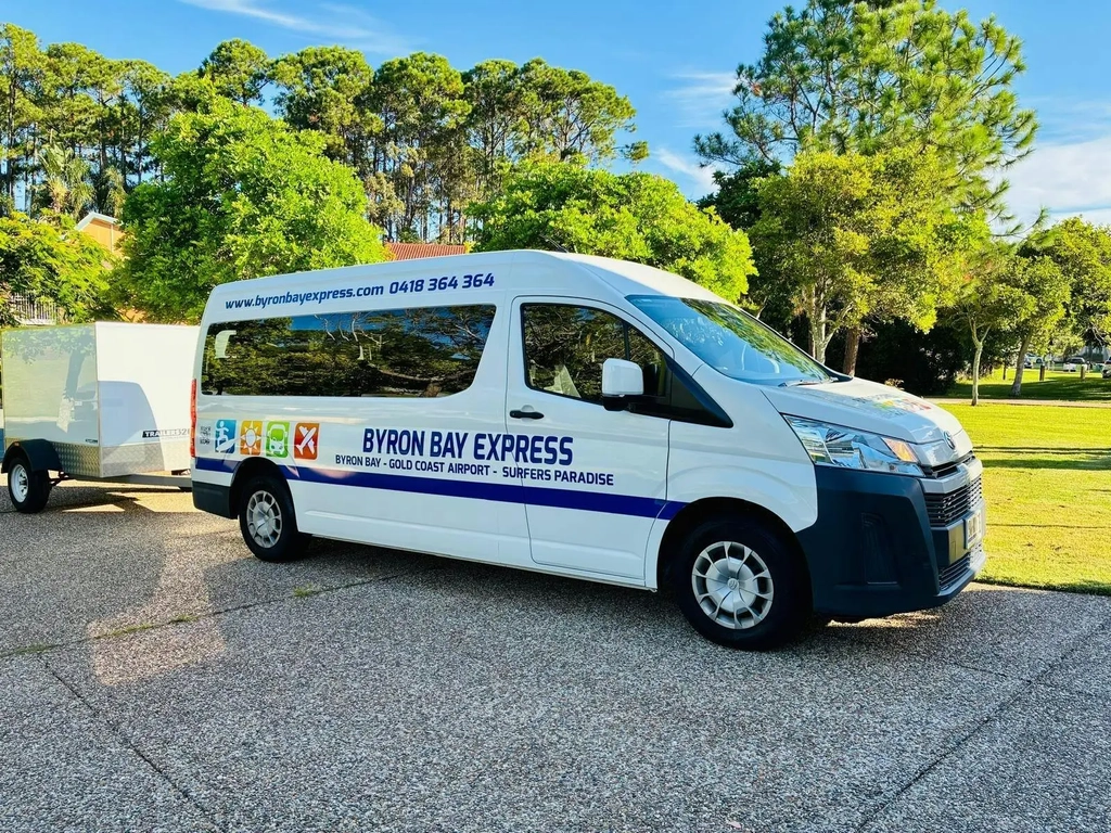 White Byron Bay Express minibus with luggage trailer parked beside trees on a sunny day