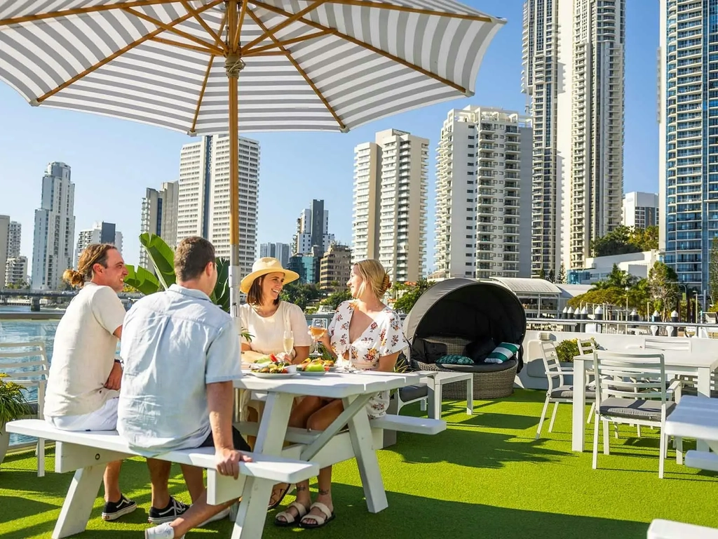 Group of four friends smiling and dining on boat sun deck with Surfers Paradise in distance.